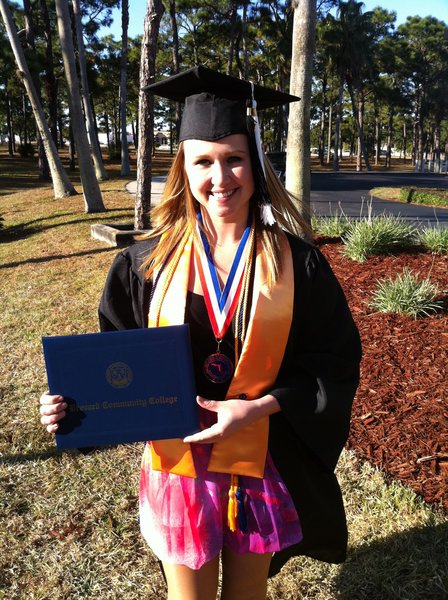 A smiling white woman with long, blonde hair (Jaime Curtis) wearing a black cap and gown, a medal, and grad regalia holds up a Brevard Community College diploma cover outdoors.