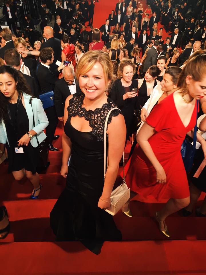 A smiling blonde woman, Jaime Braudrick, in a black dress with lace around the neckline holding a purse and standing on steps covered in red carpet within a large crowd of people in formalwear.