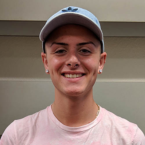 Headshot of Anthony DiBella, a smiling man with a baseball cap, studded earrings, a gold chain necklace, and a pink shirt.