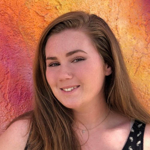 Headshot of Kristen Barg, a smiling white woman with long brown hair, a nose stud, a cross necklace, and a patterned tank top.