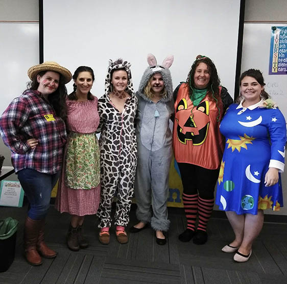 Six women in costumes (a scarecrow, a prairie woman, a giraffe, a rabbit, a pumpkin, and Miss Frizzle from "The Magic School Bus") standing at the front of a classroom with their arms around each other.