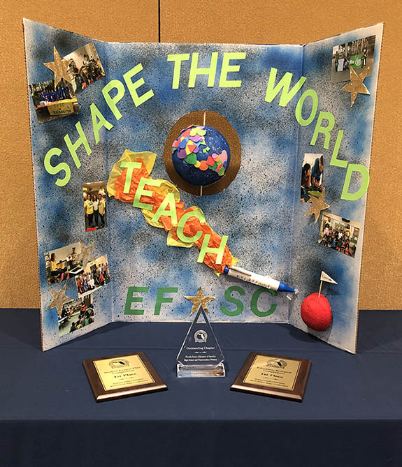 A tri-fold poster board sitting on a table with two plaques and a trophy. The poster board is covered in photos, stars, and the words, "Shape the World: Teach EFSC."
