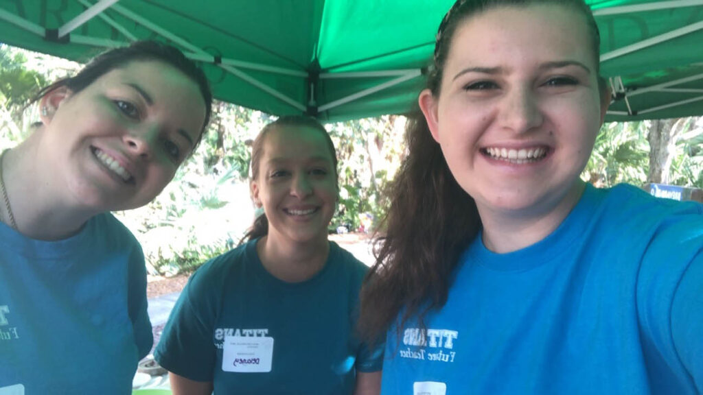 Three smiling white women with their hair pulled back and wearing Titan Future Teacher T-shirts standing under a pop-up canopy.