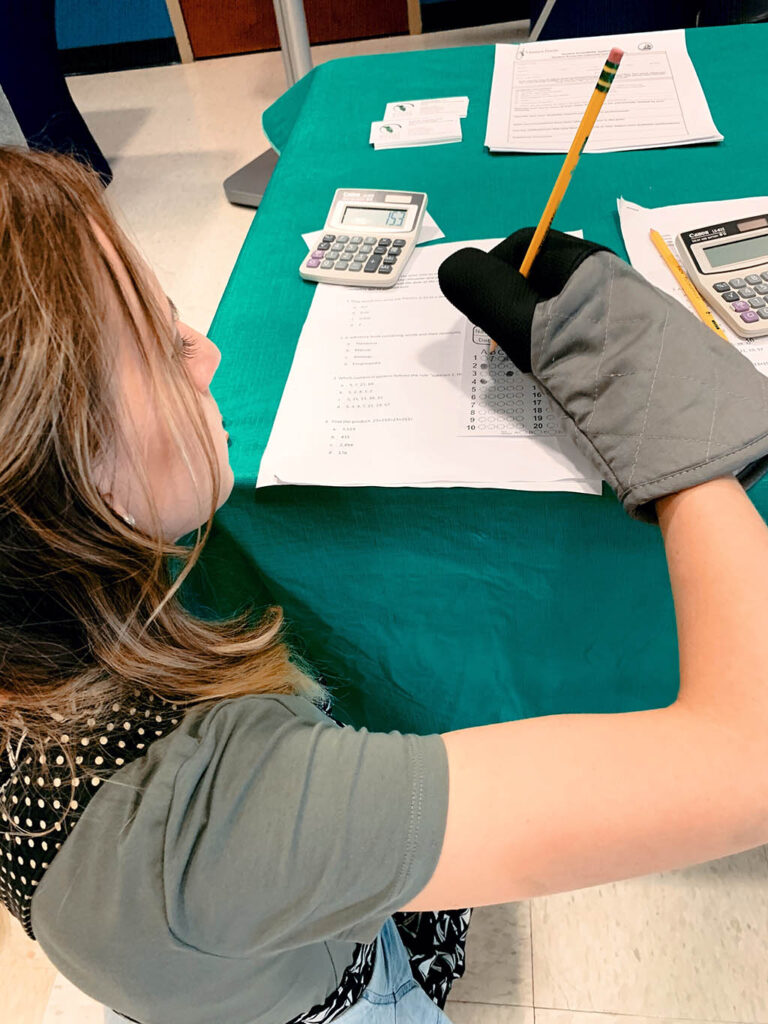 A white woman with long, dirty blonde hair (Ashley Halcom) attempts to write on paper with a pencil while wearing an oven mitt.