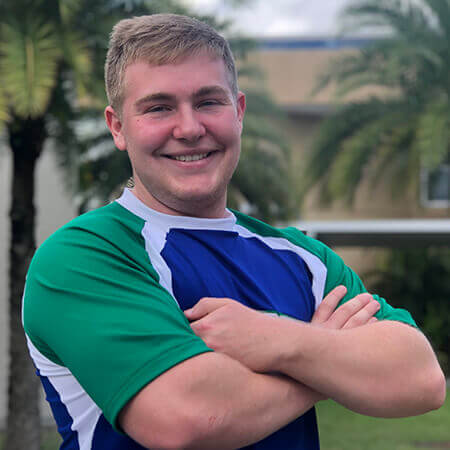 A smiling white man with blond hair, Austin Welch, shown from the chest up, wearing a blue and green cheerleading top and crossing his arms in front of a background of palm trees outside.