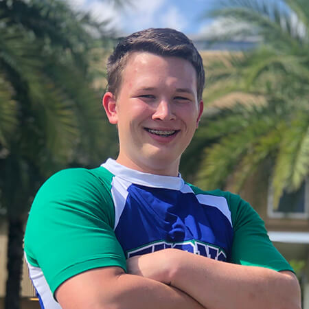 A smiling white man with light brown hair, Marcus Mercier, shown from the chest up, wearing a blue and green cheerleading top and crossing his arms in front of a background of palm trees outside.