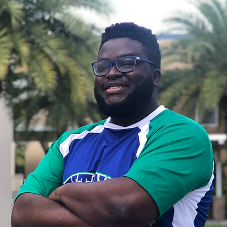 A smiling Black man with glasses, Phil Noel, shown from the chest up, wearing a blue and green cheerleading top and crossing his arms in front of a background of palm trees outside.