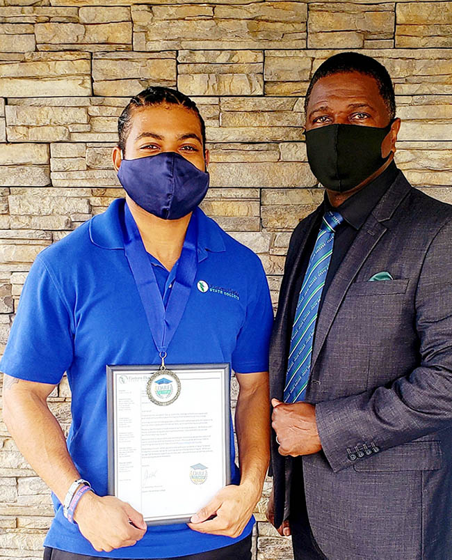 Two Black men standing in front of a brick wall: Xavier Edwards, who wears a face mask, a blue EFSC polo, and a Dare to Finish medal, holds up a framed certificate. Next to him is Michael Cadore, who wears a face mask and a suit with an EFSC tie.