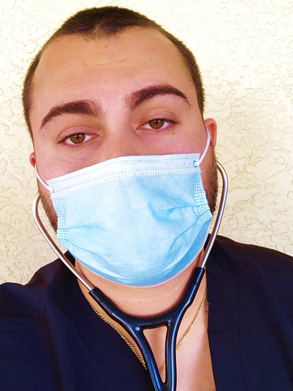 A man with short hair and a gold chain necklace shown from the chest up wearing a face mask, a stethoscope, and dark scrubs.
