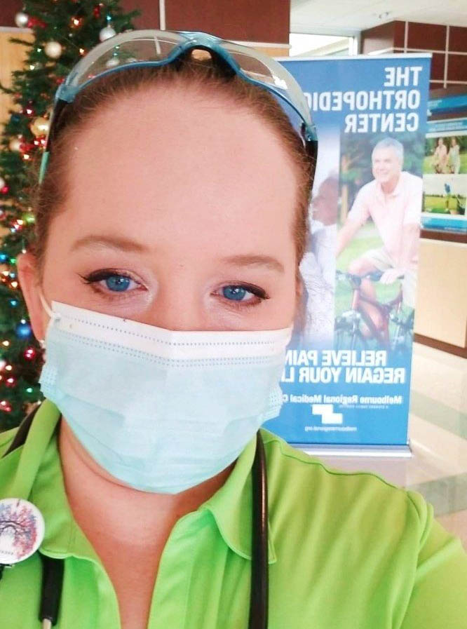 A blonde woman (Sarah Pace) in a lime green shirt with safety glasses on her head, a stethoscope around her neck, and a blue face mask stands in front of a sign and a Christmas tree.