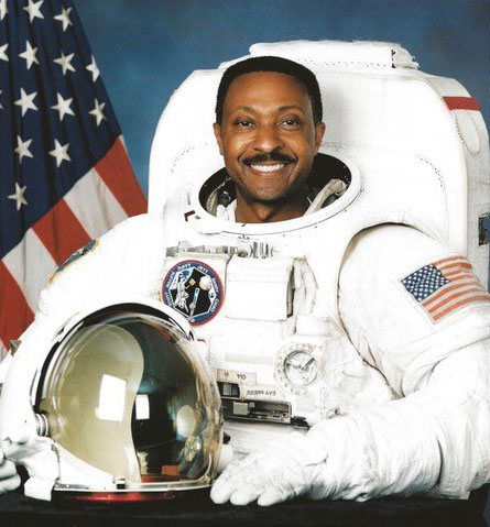 A smiling Black man, Winston Scott, sitting in front of an American flag and wearing a space suit, with the helmet resting on the surface in front of him.