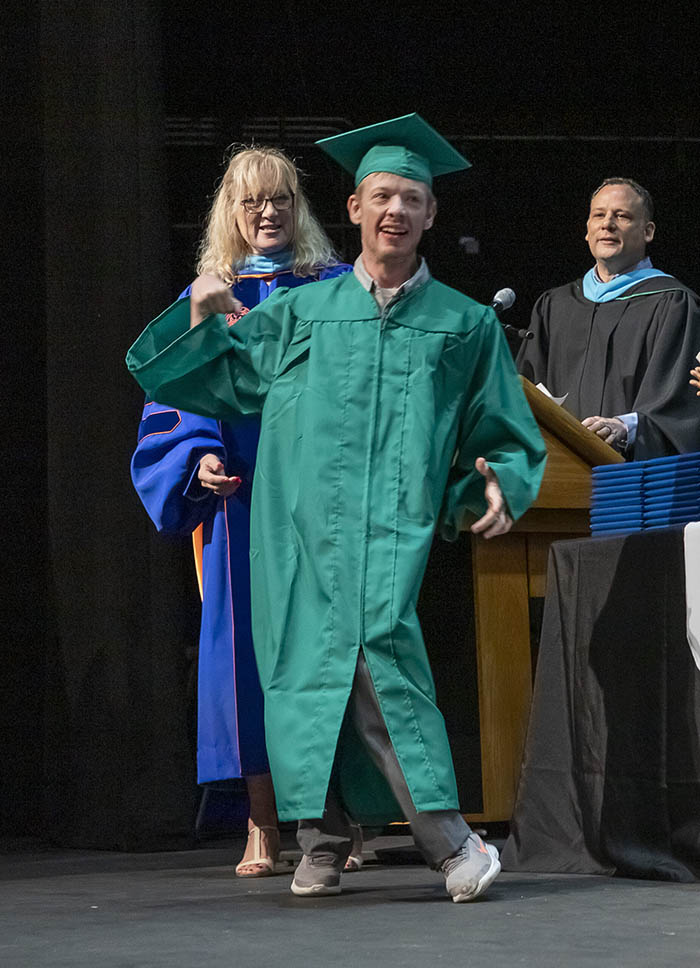 A man in a green EFSC cap and gown, Justin Miller, walks across the King Center stage, with a man and woman in robes behind him facilitating the graduation.