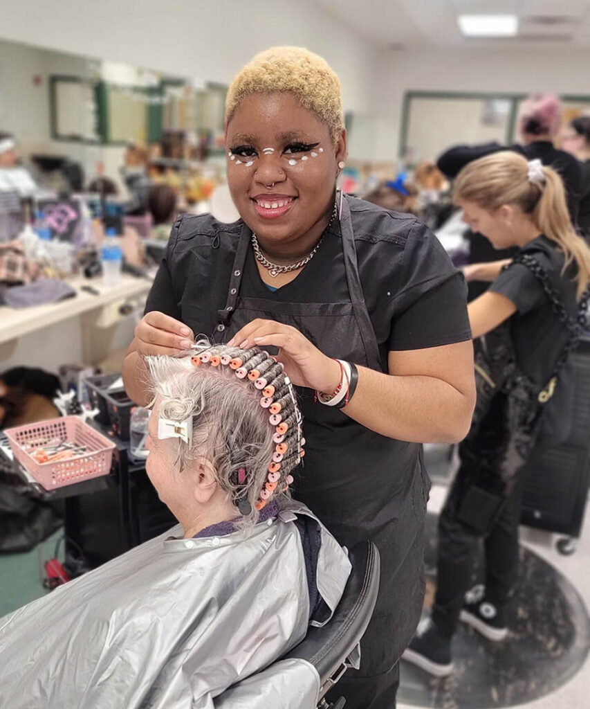A Black woman with short blonde hair, a nose ring, and white makeup under her eyes (Irie Sutton) puts curlers into an older woman's hair in the EFSC salon.
