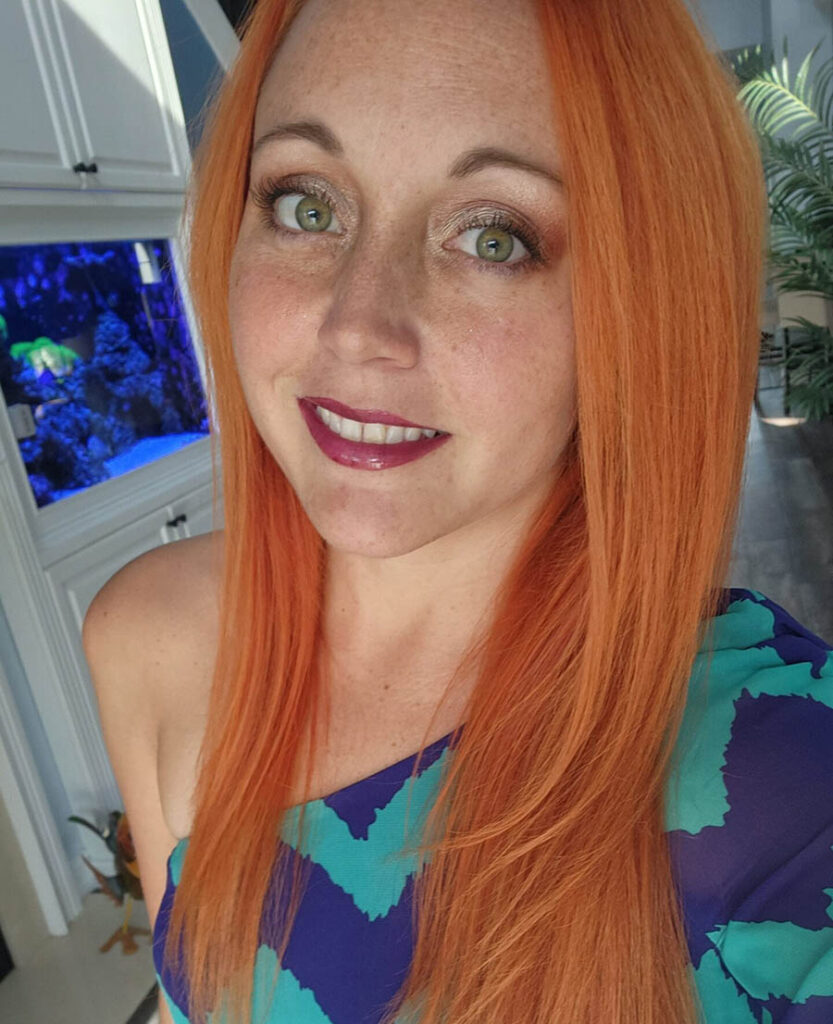 A selfie of Natasha Trythall, a redheaded woman with pink lipstick and an off-the-shoulder blue and green top.