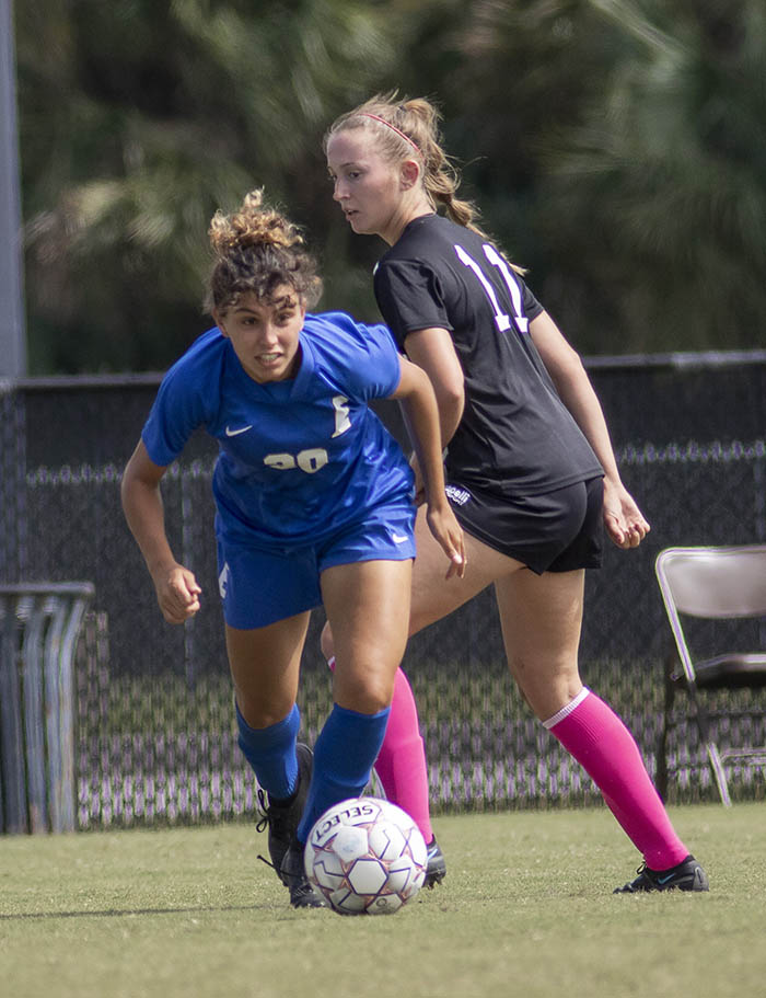 A woman (Akatarinha Papagiorgio) in a blue EFSCC Titans uniform, with her dark hair pulled up dribbles a soccer ball down a field away from a blonde woman in a black uniform.