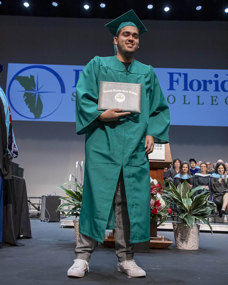 A smiling man in a green EFSC cap and gown, Joshua Katz, holds up an Eastern Florida State College diploma cover while standing on the King Center stage at graduation.
