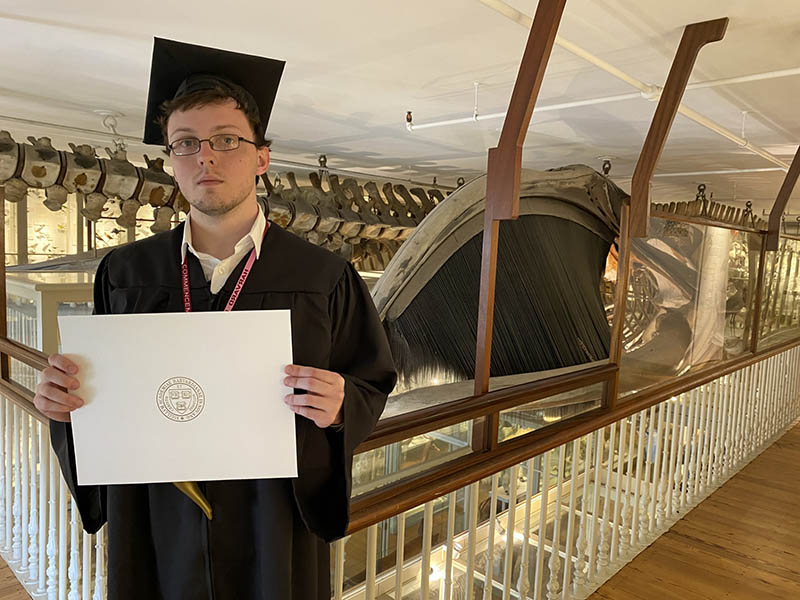 A white man in glasses and a black cap and gown, Kendall Rountree, holds up a diploma in front of a museum exhibit.
