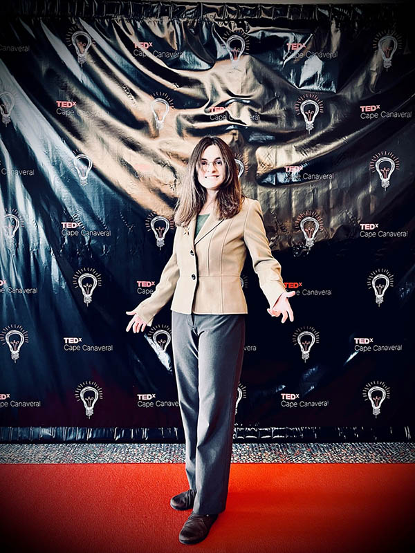 A white woman with glasses, a tan blazer, and grey slacks, Casey Covel, stands on a red carpet in front of a banner with lightbulbs and TEDx Cape Canaveral on it.