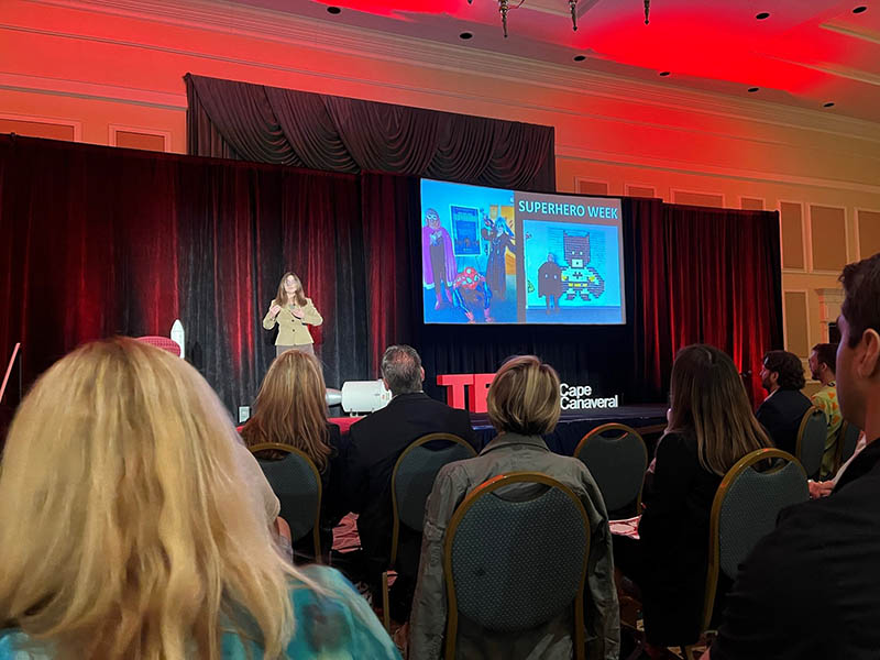 A white woman with glasses, a tan blazer, and grey slacks, Casey Covel, presents to a room full of people from a stage. A TV screen beside her displays photos from Superhero Week.