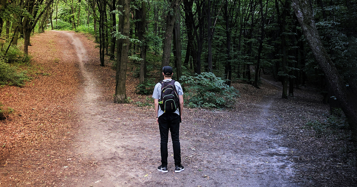 A man wearing a backpack and a hat at a crossroads in the forest. The left side is bright and the right side is dark. symbolizing the decision between A.A. vs. A.S. degrees at EFSC.