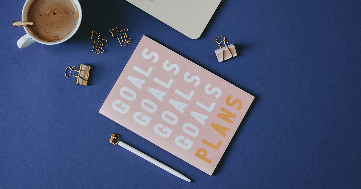 A pink notebook with "goals goals goals goals plans" written on it. Around the notebook are a pen, clips and a cup of coffee. Every item has a gold accent, representing advice from EFSC students.