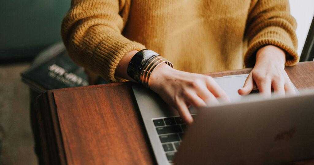 A woman in a yellow sweater typing on a laptop. She's taking advantage of the Assistive Technology Guide.