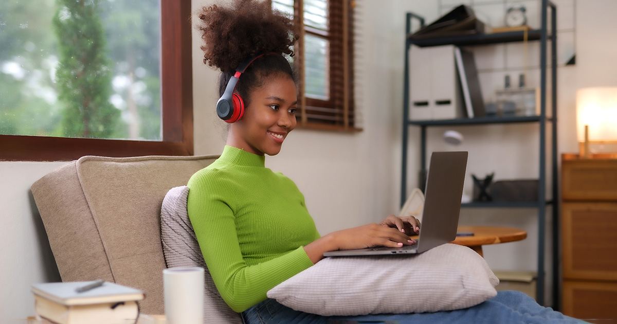 Young African American woman wearing headphones using laptop sitting on sofa in the living room and holding coffee, studying. She may have taken advantage of brain-boosting tea options to ensure effectiveness and alertness.