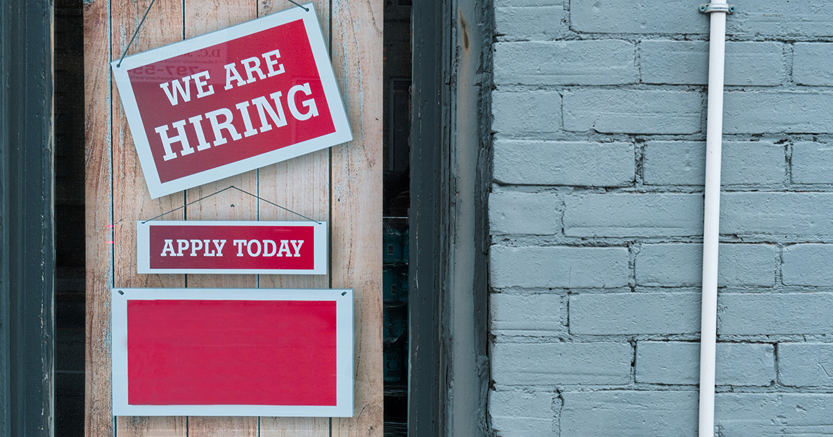 Two red signs hanging from a door. The signs have white letters that say "We Are Hiring" and "Apply Today". There is a blue brick wall next to the door. This symbolizes the hiring process and brings to mind busting workforce myths.