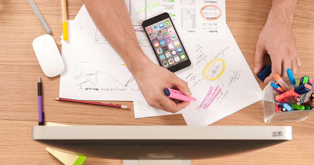 A person working at a desk highlighting scribbles on a piece of paper. Other papers, a computer and smart phone are scattered on the desk. It evokes thoughts of choosing a college major.