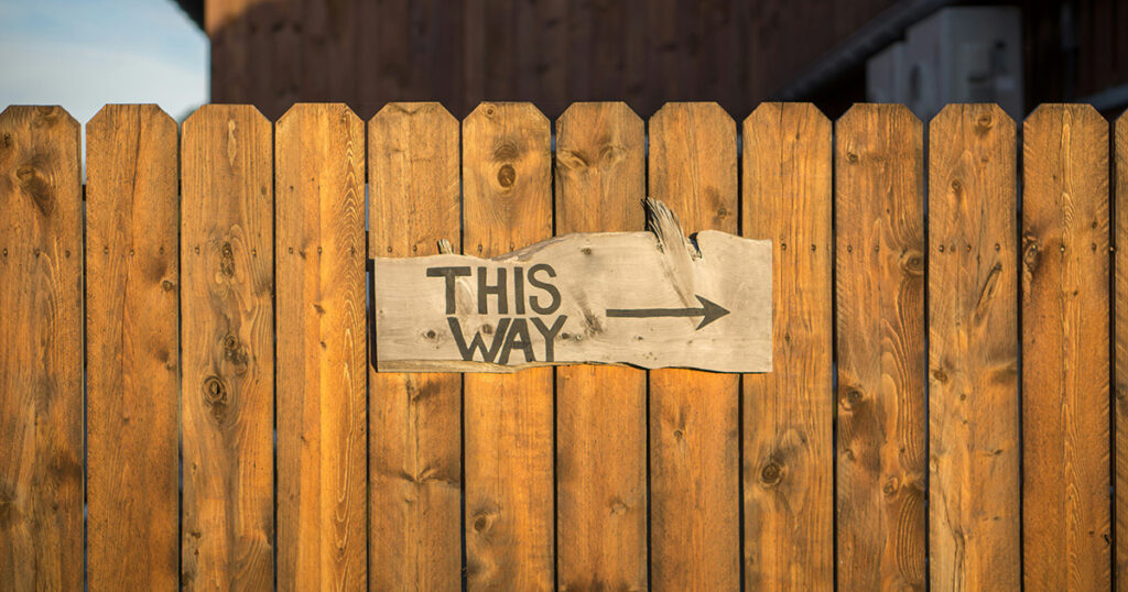 A brown fence with a rustic grey sign on it. The sign has black writing that says "This way" and has an arrow pointing right. The images serves as a metaphor for the college admissions guide.