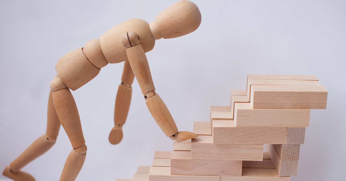 A wooden articulated figure bending over a wooden staircase as though struggling to climb it. The image serves as a metaphor for college freshman advice given to incoming students.