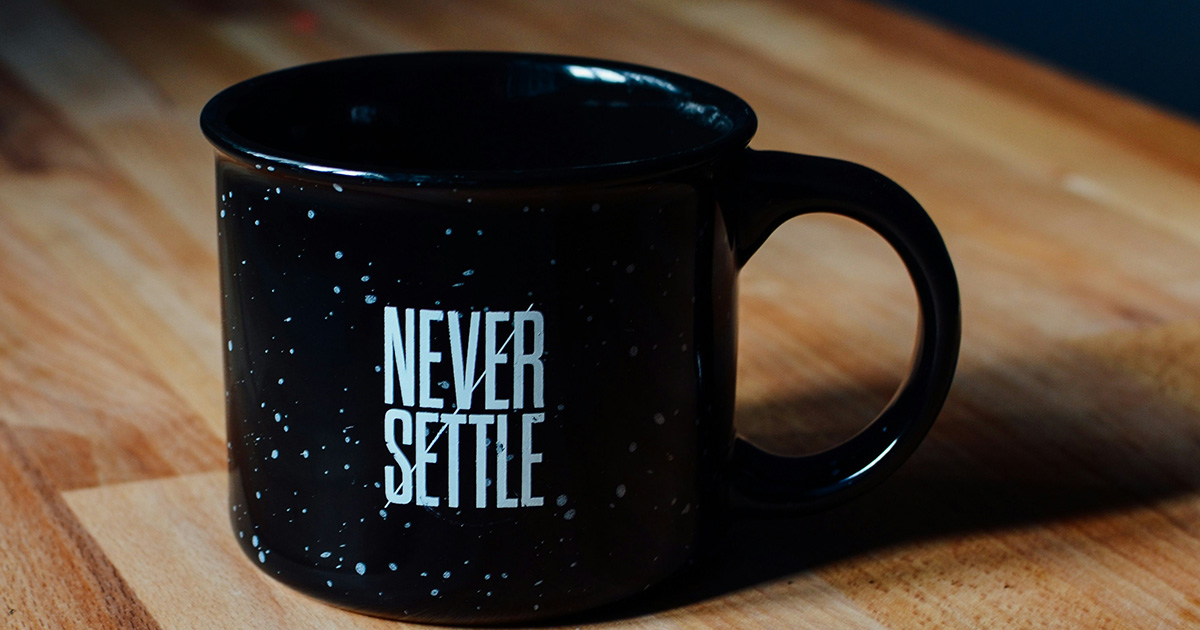 A black mug with "NEVER SETTLE" written on it. This mantra represents college motivation reasons that students may use to persevere through their degree program.