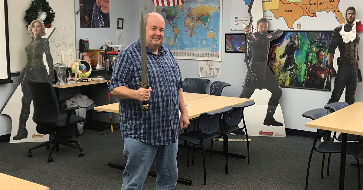 Luke Leonard holding a prop sword in his classroom. There are Marvel Avenger cardboard cut outs in the background