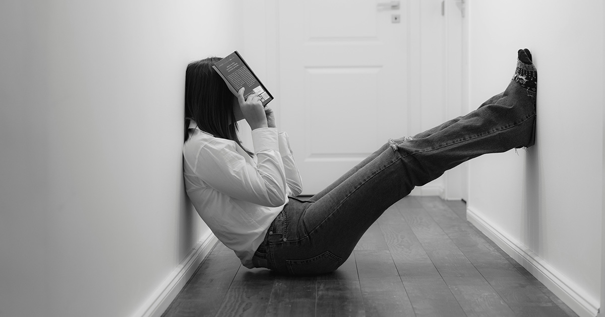 A woman sitting in a small hallway with back to one wall and her legs propped up on the other. She is covering her face with a book, trying to master effective study tips.