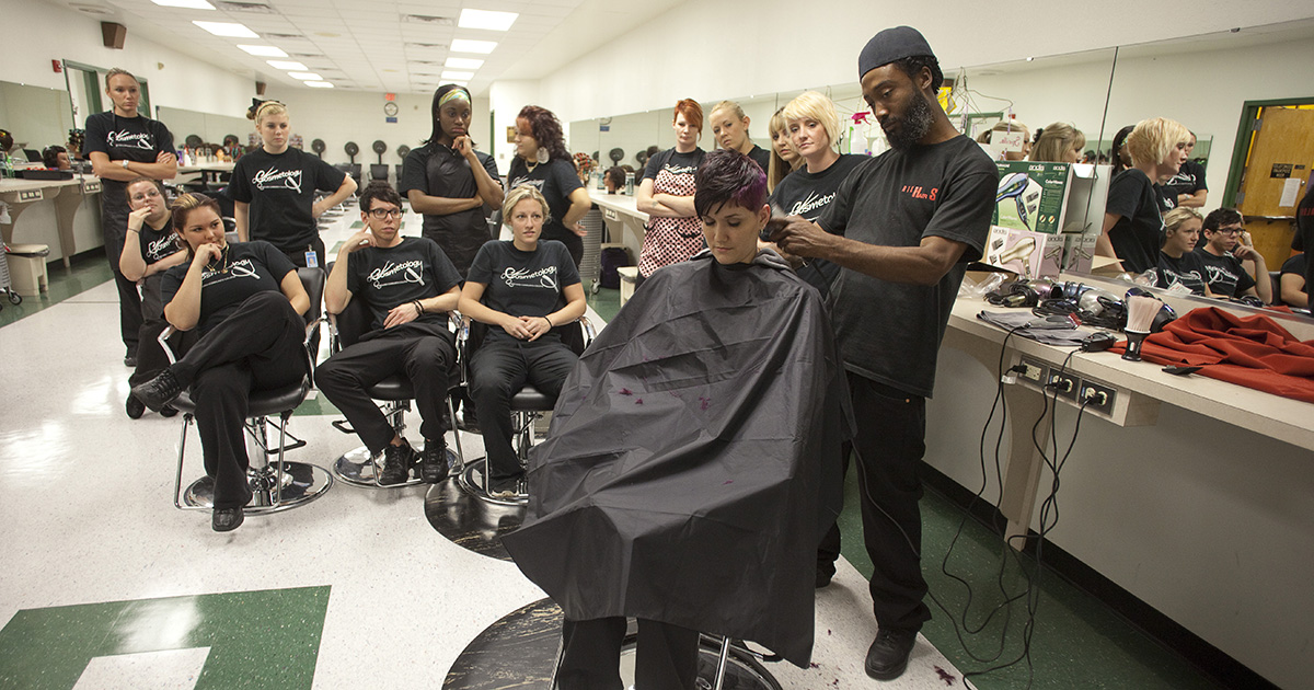 A male cosmetologist in EFSC's cosmetology program working cutting the hair of a woman in a chair. A group of cosmetology students are in the background watching.