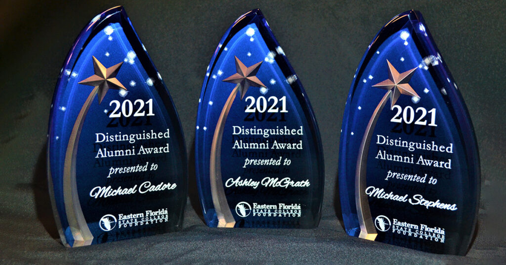 Three blue triangular award statues representing the EFSC Distinguished Alumni Award. The awards have the text "2021 Distinguished Alumni Award presented to Michael Cadore, Ashley McGrath, and Michael Stephens".