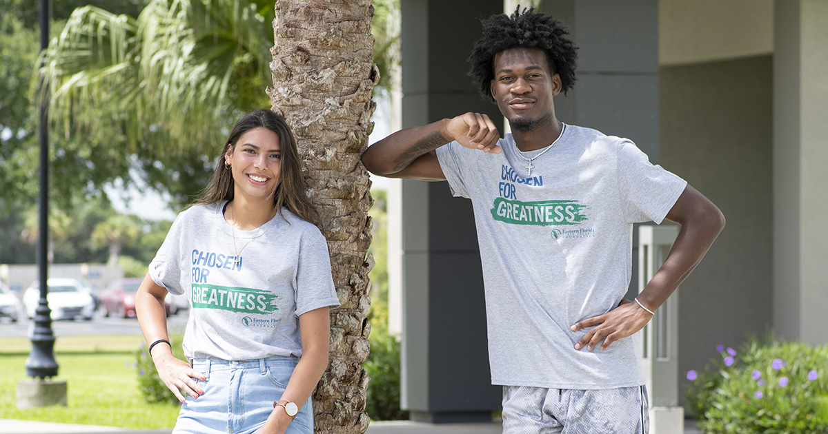 Two Eastern Florida State College students, a Hispanic female and a Black male, stand smiling and posing next to a palm tree. They are both wearing shirts that say "Chosen for Greatness". The image represents the process of EFSC Scholarship Fundraising.