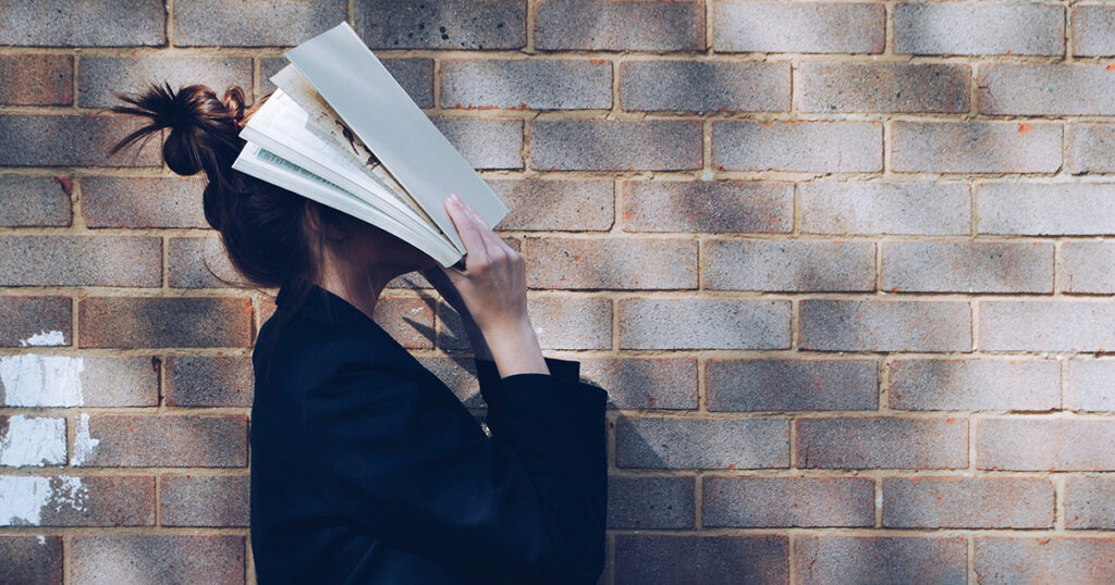 A woman covering her face with a book. A brick wall is in the background. The image is a metaphor for FAFSA Financial Aid Standards.