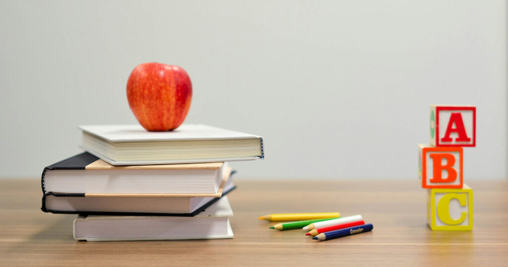 A stack of four books with an apple on top sitting on a desk. Some colored pencils and a stack of "ABC" blocks sit next to the books, representing first-year teacher advice.