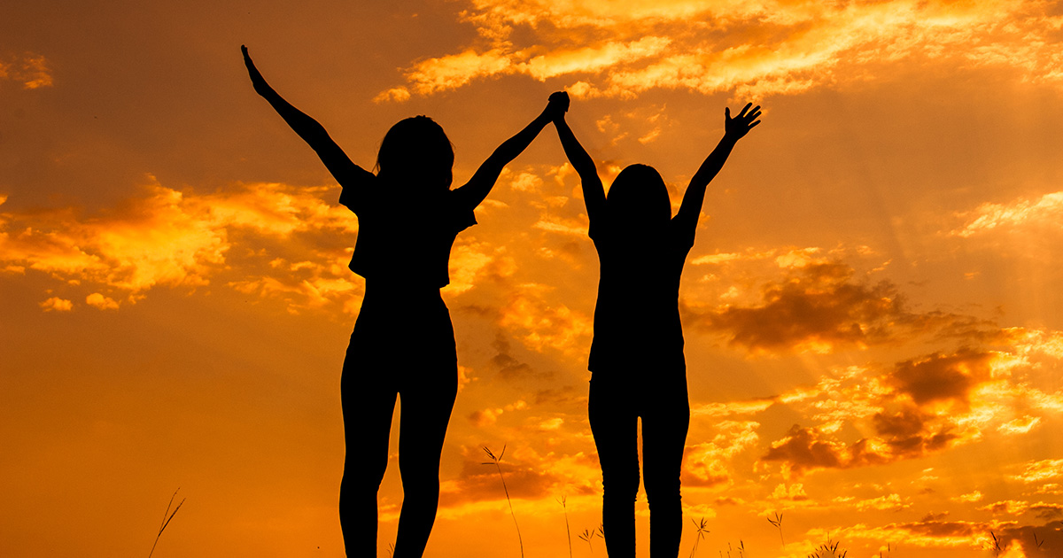 Two women holding hands with arms raised looking towards a sunset. It symbolizes the victory attained over freshman burnout.