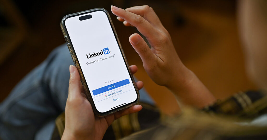 A female using her smartphone, holding a smartphone with LinkedIn logo on screen. She is preparing for LinkedIn profile optimization.