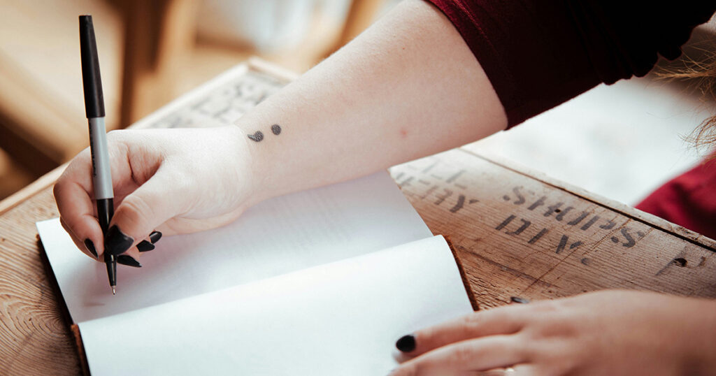 A person with a semi-colon tattoo on their wrist writing in a journal, representing mental health in college.