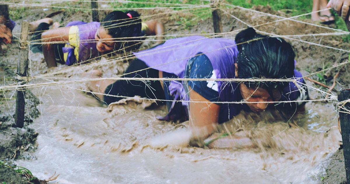 Women crawling through the mud under string in an obstacle course. She's implementing overcoming challenges tips also beneficial for college.