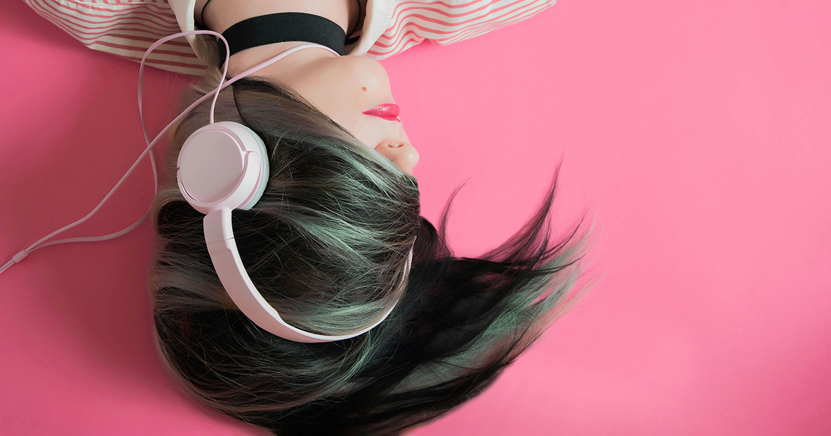 A woman lying down with her black and silver hair covering her eyes. She is wearing pink headphones and has on pink lipstick. The background is pink. The image symbolizes playlists every student needs.