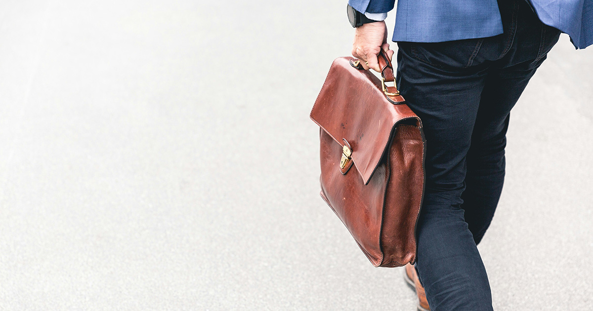 A man wearing blue pants, a blue jacket, and a watch walking holding a brown leather briefcase. The image symbolizes resume writing mistakes the man may have made prior to his job application.