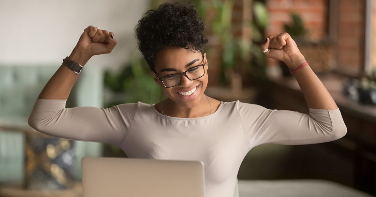 Excited African American woman rejoicing with arms up in front of laptop. She's successfully used scholarship essay writing tips to write a winning scholarship essay.