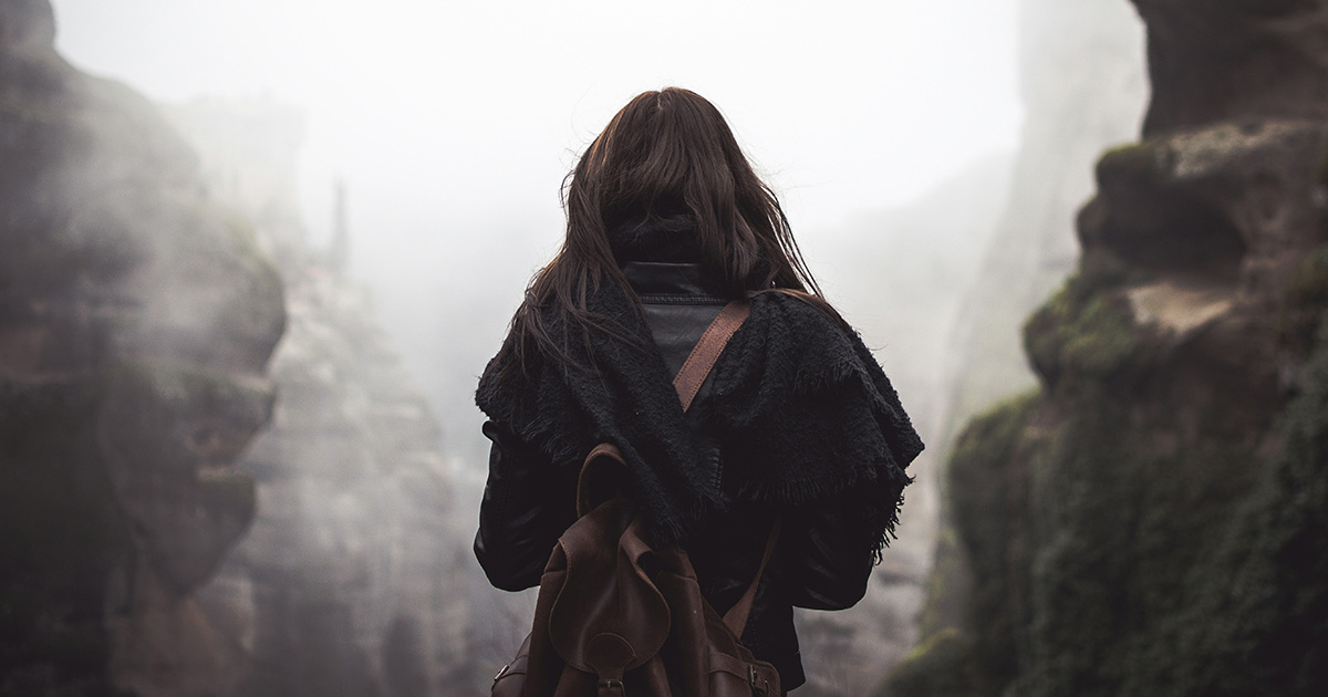 The back of a woman with brown hair, a black scarf, and a brown leather backpack. She's walking through fog with cliffs on either side of her, symbolizing tutoring session preparation tips.