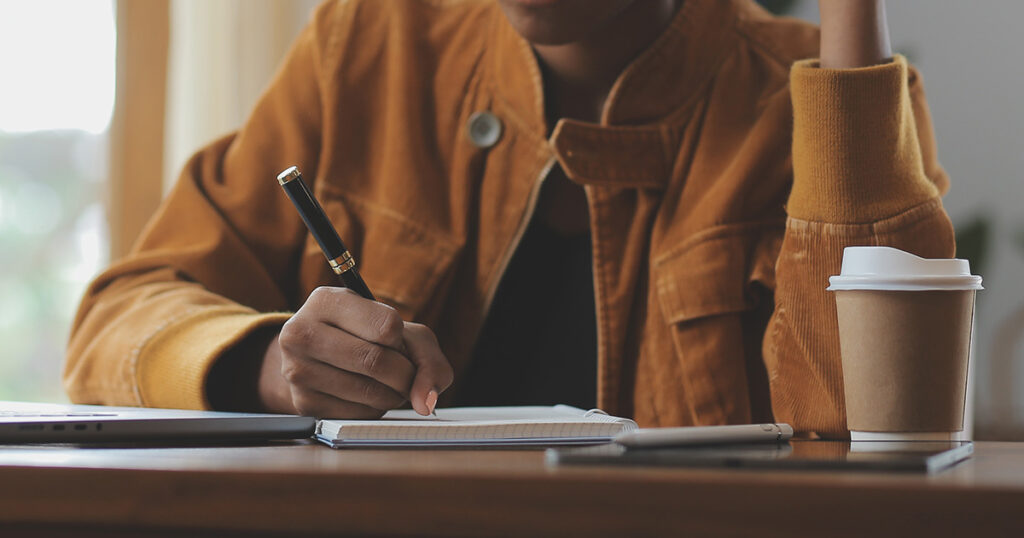 A Latina woman writing in a notebook next to a laptop and cup of coffee. She may be using unconventional studying hacks to help her master the material.