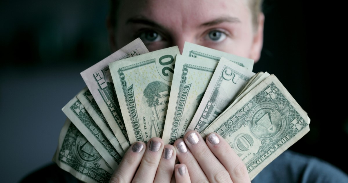 A person holding different denominations of USD in front of their face. All of their face is obscured by the money except their eyes, as they ask you to contemplate the value of education.