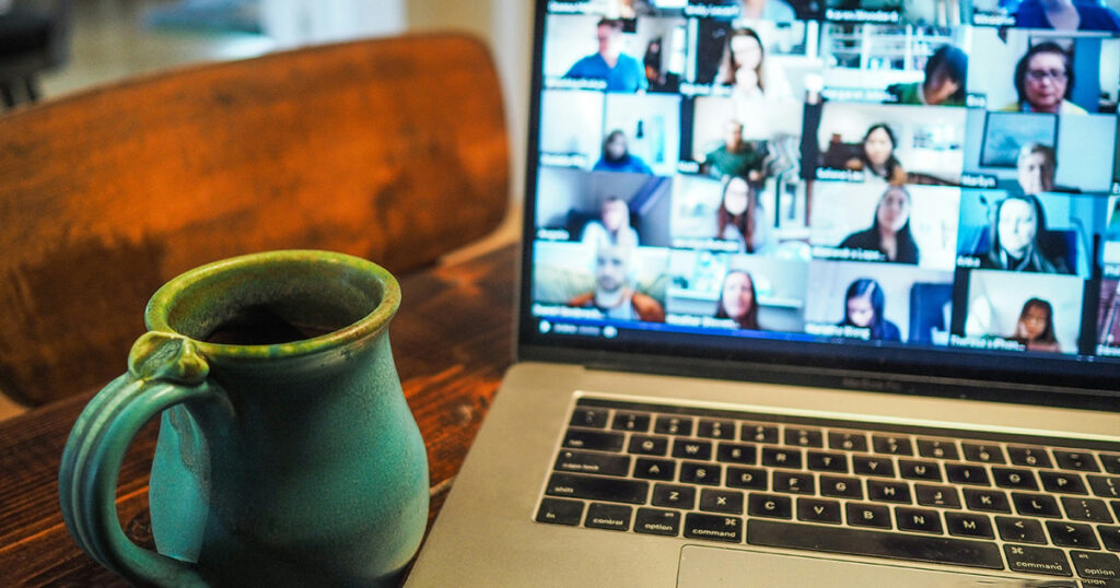 A laptop on a table. The screen of the laptop is showing a zoom call with many participants. There is a green mug on the table next to the laptop. It calls to mind the fact that virtual has become reality.